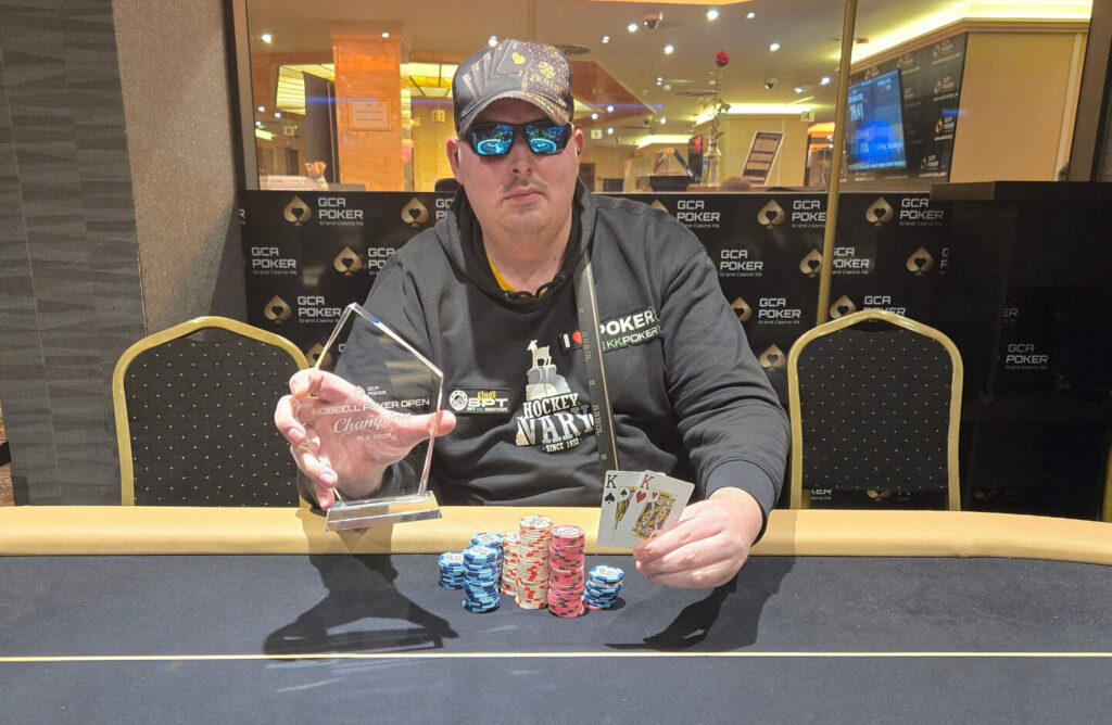 Ein eleganter Mann in einem noblen Anzug sitzt lässig an einem Pokertisch und hält eine Hand mit Spielkarten. Im Hintergrund sind andere Spieler und eine glamouröse Atmosphäre zu erkennen.