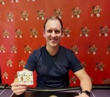 Ein Mann in einem schwarzen Anzug und mit Sonnenbrille sitzt an einem Pokertisch und hält zwei Karten in der Hand, während er konzentriert auf seine Gegner schaut.