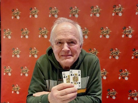 Ein Mann mit dunklen Haaren und einem gestreiften Hemd sitzt an einem Pokertisch und hält Karten in der Hand. Er schaut konzentriert auf seine Karten.