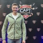 Ein Mann sitzt an einem Pokertisch und hält zwei Spielkarten in der Hand, während er konzentriert auf seine Gegner schaut. In der Mitte des Tisches liegen bereits mehrere Karten. Die Atmos