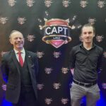 Ein Mann sitzt an einem Pokertisch und hält zwei Spielkarten in der Hand. Er schaut konzentriert auf den Tisch, auf dem Pokerchips und weitere Karten liegen. Im Hintergrund sind weitere