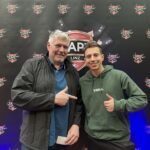 Ein Mann und eine Frau sitzen an einem Pokertisch und spielen Poker. Der Mann hat eine Hand mit Chips in der Luft und die Frau schaut konzentriert auf ihre Karten. Im Hintergrund sind Pokerchips und Spiel