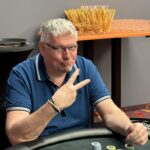 Ein Mann sitzt an einem Pokertisch und hält zwei Spielkarten in der Hand. Er trägt eine schwarze Kappe und ein dunkles Hemd. Auf dem Tisch liegen Chips und Karten. Im Hintergrund