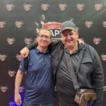 Ein Mann und eine Frau sitzen an einem Pokertisch. Der Mann hält Karten in der Hand, während die Frau konzentriert auf ihre Chips schaut. Hinter ihnen sind leuchtende Spielautomaten zu sehen.