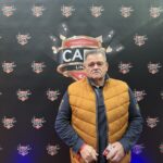 Ein Mann sitzt an einem Pokertisch und hält eine Hand mit Spielkarten. Er trägt eine dunkle Brille und konzentriert sich auf sein Blatt. Im Hintergrund sind andere Spieler und Chips zu sehen.