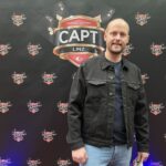 Ein Mann sitzt an einem Pokertisch und hält zwei Karten in der Hand, während er konzentriert auf die Spielkarten auf dem Tisch blickt. Im Hintergrund sind weitere Spieler und Pokerchips zu