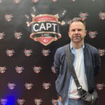 Ein glücklicher Mann sitzt an einem Pokertisch und hält eine Hand mit Karten. Er lächelt und schaut konzentriert auf seine Hand. Im Hintergrund sind Pokerchips und andere Spieler zu sehen.