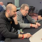 Ein Mann sitzt an einem Pokertisch, hält Karten in der Hand und schaut konzentriert auf seine Gegner. Im Hintergrund sind Chips und Getränke zu sehen.