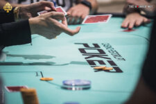 Ein Mann sitzt an einem Pokertisch und hält zwei Karten in der Hand. Er schaut konzentriert auf die Gemeinschaftskarten auf dem Tisch. Im Hintergrund sind weitere Spieler und Dealer zu sehen,