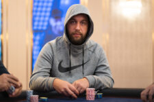 Ein junger Mann sitzt an einem Pokertisch in einem Casino, umgeben von anderen Spielern. Er hält seine Karten in der Hand und schaut konzentriert auf den Tisch. Eine Rauchwolke ste