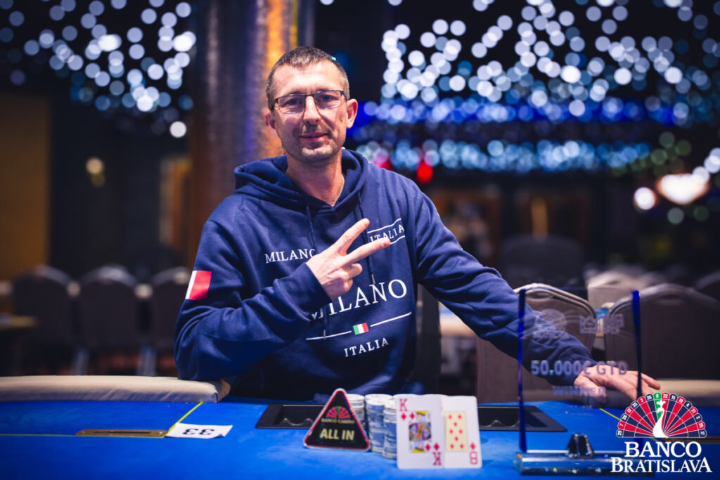 Ein Mann, der eine schwarze Sonnenbrille trägt, sitzt an einem Pokertisch und hält zwei Spielkarten in der Hand. Im Hintergrund sind andere Spieler und Pokerchips zu sehen.