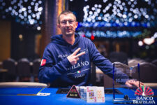 Ein Mann, der eine schwarze Sonnenbrille trägt, sitzt an einem Pokertisch und hält zwei Spielkarten in der Hand. Im Hintergrund sind andere Spieler und Pokerchips zu sehen.