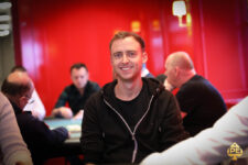 Ein Mann namens Alex Tkatschew sitzt lächelnd an einem Pokertisch und hält Karten in der Hand. Er trägt ein dunkles Hemd und eine Sonnenbrille. Im Hintergrund sind weitere