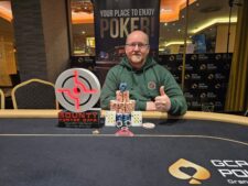 Ein Mann in einem schwarzen Anzug und einer schwarzen Sonnenbrille hält eine Hand mit Pokerchips und schaut ernst in die Kamera.