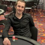Ein Mann sitzt an einem Pokertisch mit Chips und Karten vor sich. Er konzentriert sich auf sein Blatt und hat eine ernste Miene.