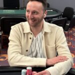 Ein Mann sitzt an einem Pokertisch und hält zwei Spielkarten in der Hand. Er hat eine ernste Miene und scheint konzentriert zu sein. Um ihn herum sitzen mehrere Personen, die ebenfalls K