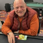 Ein Gruppenfoto von fünf Pokerspielern, die konzentriert an einem Tisch sitzen und Karten halten. Die Spieler haben unterschiedliche Gesichtsausdrücke und tragen Kopfhörer.