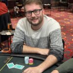 Ein professioneller Pokerspieler mit Sonnenbrille und Kappe, der konzentriert Karten auf einem grünen Pokertisch betrachtet.