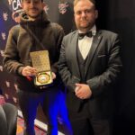 Ein Mann in einem schwarzen Anzug und einer Sonnenbrille sitzt lässig an einem Pokertisch und hält Pokerkarten in der Hand, während er ernsthaft auf seine Gegner schaut.