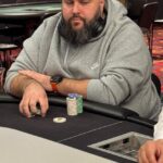 Ein professioneller Pokerspieler in Aktion während eines Turniers, umgeben von anderen Spielern und Zuschauern in einem gut beleuchteten Veranstaltungsraum.