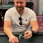 Ein professioneller Pokerspieler sitzt an einem Tisch während eines Turniers und hält seine Karten verdeckt in der Hand. Die Spannung ist spürbar, während er seine Mitspieler beobachtet und über