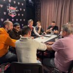 Ein professioneller Pokertisch mit grüner Filzoberfläche, umgeben von Spielern in eleganten Anzügen und mit Pokerchips vor sich. Ein Dealer mischt Karten in der Mitte des Tisches.