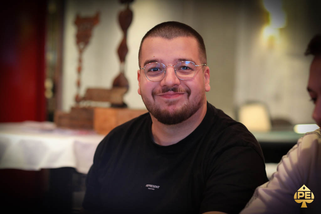 Ein Mann mit dunklen Haaren und Bart, der ein dunkles Hemd trägt und lächelnd in die Kamera schaut.