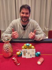 Ein Mann und eine Frau sitzen am Pokertisch und lächeln sich an, während sie ihre Karten betrachten. Die Frau hält eine Hand mit einem hohen Wert, während der Mann seine Karten mit einem überraschten