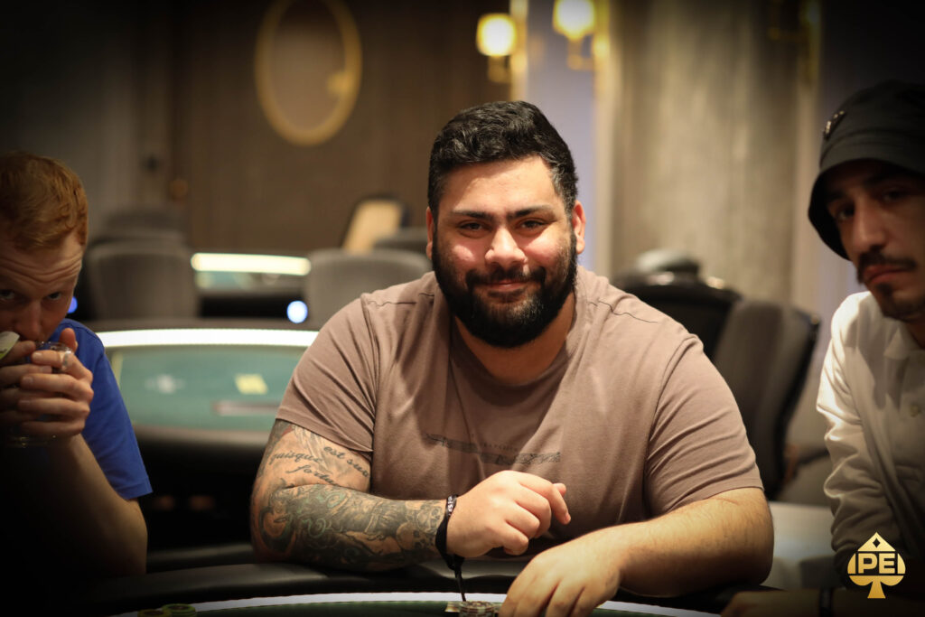 Ein Mann sitzt an einem Pokertisch und hält zwei Spielkarten in der Hand. Er hat dunkles Haar und trägt ein schwarzes Hemd. Im Hintergrund sind Pokerchips und andere Spieler zu sehen.
