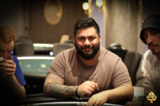 Ein Mann sitzt an einem Pokertisch und hält zwei Spielkarten in der Hand. Er hat dunkles Haar und trägt ein schwarzes Hemd. Im Hintergrund sind Pokerchips und andere Spieler zu sehen.