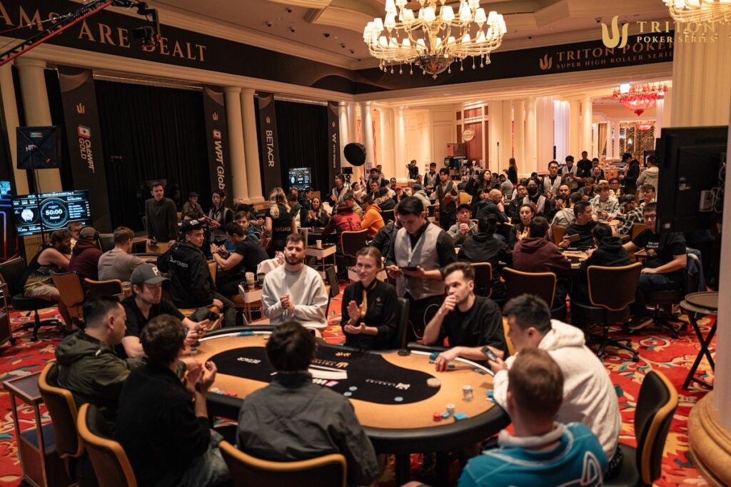 Ein Foto von einer großen Pokertischrunde bei einem Triton Jeju Event. Mehrere Spieler sitzen um den Tisch herum, Karten und Chips liegen auf dem Tisch. In der Mitte des Tisches befindet