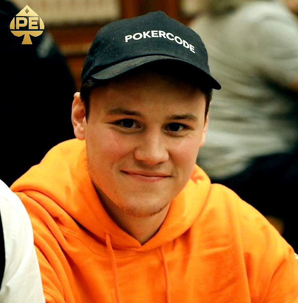Ein Mann mit dunklen Haaren und einem Bart sitzt an einem Pokertisch und hält Karten in der Hand. Er trägt ein schwarzes Hemd und eine schwarze Kappe. Im Hintergrund sind weitere Pokerspi