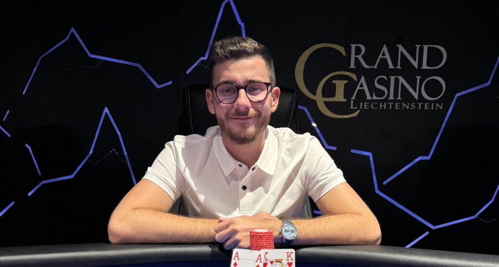 Ein Mann und eine Frau sitzen an einem Pokertisch und spielen mit Pokerchips und Karten. Der Mann hat einen ernsten Gesichtsausdruck, während die Frau lächelt. Im Hintergrund sind weitere Pokert