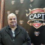 Ein professioneller Pokertisch mit grünem Filz, umgeben von Spielkarten, Pokerchips und Getränken. Mehrere Personen sitzen mit ernstem Gesichtsausdruck um den Tisch herum und