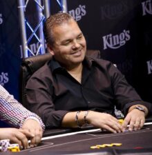 Ein Mann mit dunklen Haaren und Bart sitzt an einem Pokertisch. Er trägt ein dunkles Oberteil und hat eine Sonnenbrille auf der Stirn. In seiner Hand hält er Pokerchips.