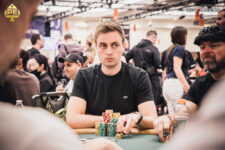 Ein Mann namens Leonard Maue sitzt an einem Pokertisch bei der World Series of Poker Main Event. Er trägt eine dunkle Brille und konzentriert sich auf seine Karten. Andere Spieler und Zuschauer sind