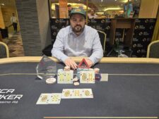 Ein Tisch mit grünem Filz, auf dem Pokerchips, Spielkarten und Getränke zu sehen sind. Vier Personen sitzen um den Tisch und spielen Poker.