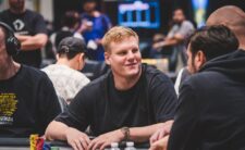 Ein junger Mann sitzt an einem Pokertisch und hält Karten in der Hand. Er trägt ein schwarzes Hemd und hat kurze dunkle Haare. Im Hintergrund sind weitere Spieler zu sehen.