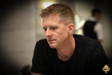 Ein junger Mann namens Maxi Lehmannski sitzt lächelnd an einem Pokertisch und hält seine Karten in der Hand. Er trägt eine dunkle Brille und ein schwarzes Hemd. Im Hinter