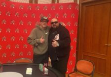 Ein Mann mit einer Sonnenbrille und einem Hut sitzt an einem Pokertisch und hält zwei Asse als Startkarten. Die anderen Spieler am Tisch sind verschwommen im Hintergrund zu sehen.