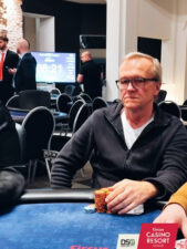 Ein älterer Mann mit grauen Haaren, der eine Brille trägt, sitzt an einem Pokertisch und betrachtet nachdenklich seine Karten. Er hat eine konzentrierte und ernste Miene.