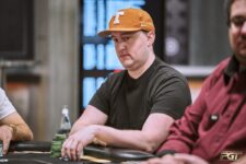 Ein Mann sitzt an einem Pokertisch während eines Turniers. Er trägt eine schwarze Kappe und konzentriert sich auf seine Karten. Andere Spieler und Dealer sind im Hintergrund zu sehen.