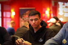Ein junger Mann mit dunklen Haaren und einem Bart sitzt am Pokertisch. Er trägt ein schwarzes Hemd und eine schwarze Brille. Seine Hand hält Spielkarten, während er konzentriert auf