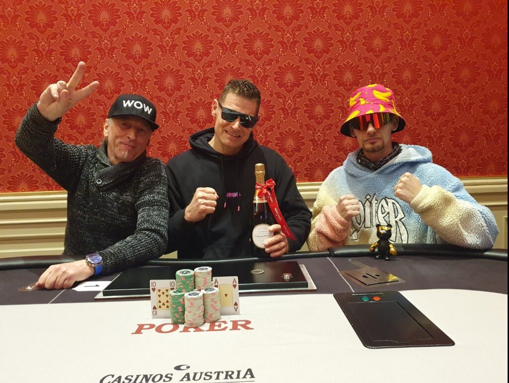 Ein Pokal mit einem Pokerchip-Design, umgeben von Spielkarten und Jetons auf einem Tisch. Im Hintergrund sind Spieler zu sehen, die sich freuen.