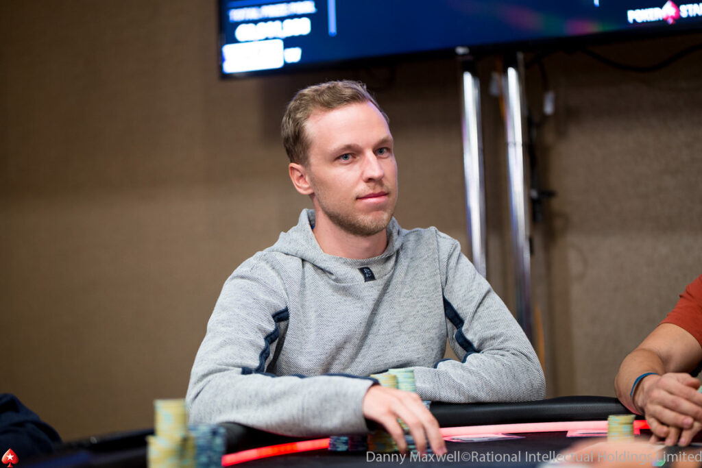 Ein junger Mann in einem dunklen Anzug sitzt an einem Pokertisch mit Chips und Karten vor sich, während er konzentriert auf sein Blatt schaut. Im Hintergrund sind andere Spieler und Dealer zu sehen.