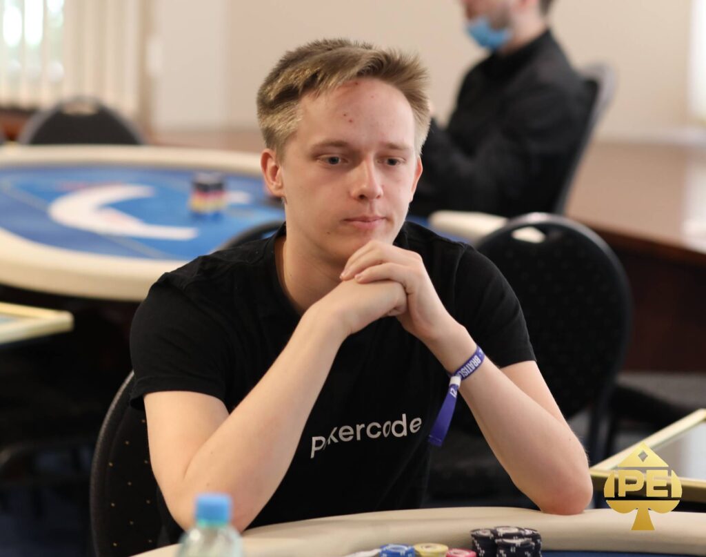 Ein Mann mit kurzen braunen Haaren und einem grauen Hemd sitzt an einem Pokertisch und hält Karten in der Hand. Er schaut konzentriert auf seine Karten und hat eine ernste Miene