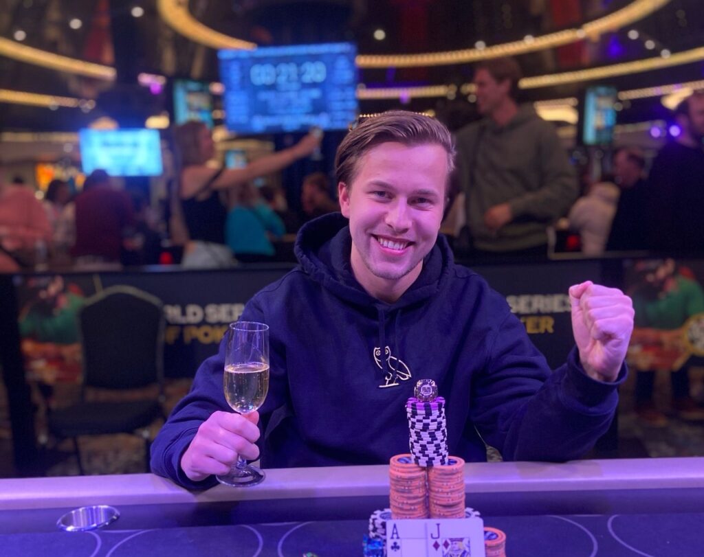 Ein Mann in einem dunklen Anzug sitzt an einem Pokertisch und hält triumphierend eine Spielkarte in der Hand. Im Hintergrund sind weitere Spieler und eine elegante Casinoumgebung zu sehen.