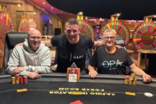 Ein Mann mit einem breiten Lächeln steht auf einem Podium, umgeben von Konfetti und jubelnden Menschen. Er hält einen großen Pokal in die Luft.
