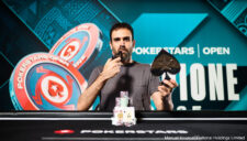 Ein Mann sitzt an einem Pokertisch und hält zwei Spielkarten in der Hand. Er trägt ein dunkles Hemd und eine Sonnenbrille. Im Hintergrund sind Pokerchips und weitere Spieler zu sehen.