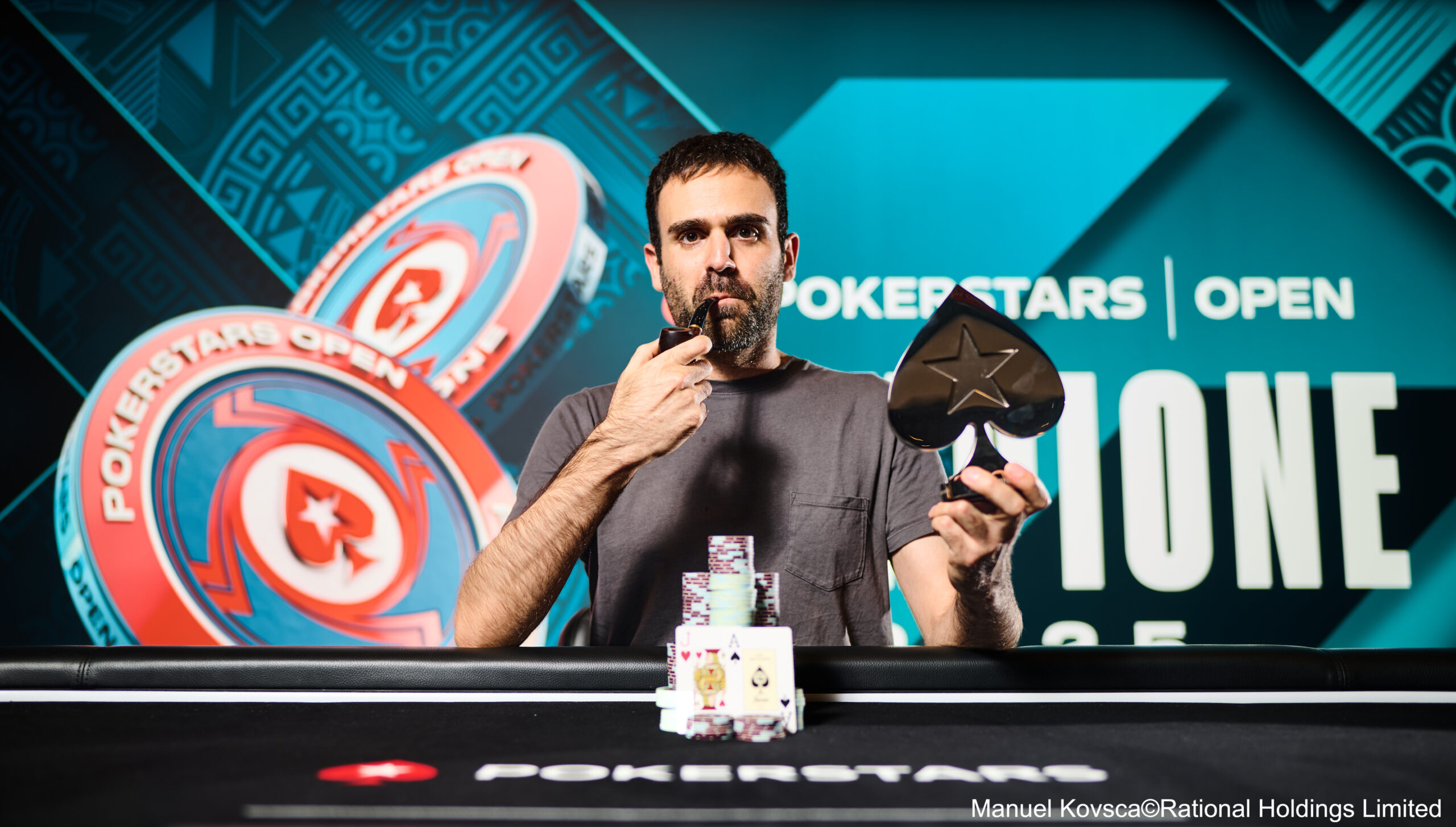 Ein Mann sitzt an einem Pokertisch und hält zwei Spielkarten in der Hand. Er trägt ein dunkles Hemd und eine Sonnenbrille. Im Hintergrund sind Pokerchips und weitere Spieler zu sehen.